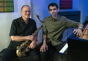 Two men pose indoors; one holds a saxophone while seated, and the other sits at a piano. Both wear collared shirts and look at the camera.