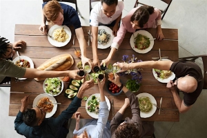 Eight people sit around a wooden table with plates of food and glasses, raising their drinks in a toast during a meal.