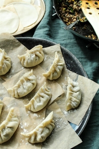 A plate of uncooked dumplings on parchment squares, with a stack of dumpling wrappers and a pan of filling in the background.