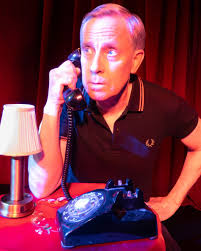A man in a black polo shirt holds a rotary phone to his ear, looking up, seated at a table with a lamp and red tablecloth.