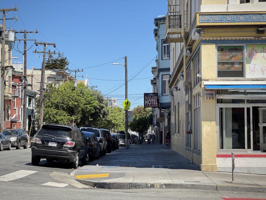 A city street with parked cars, a yellow building with a corner store, and a hotel sign visible in the background on a sunny day.