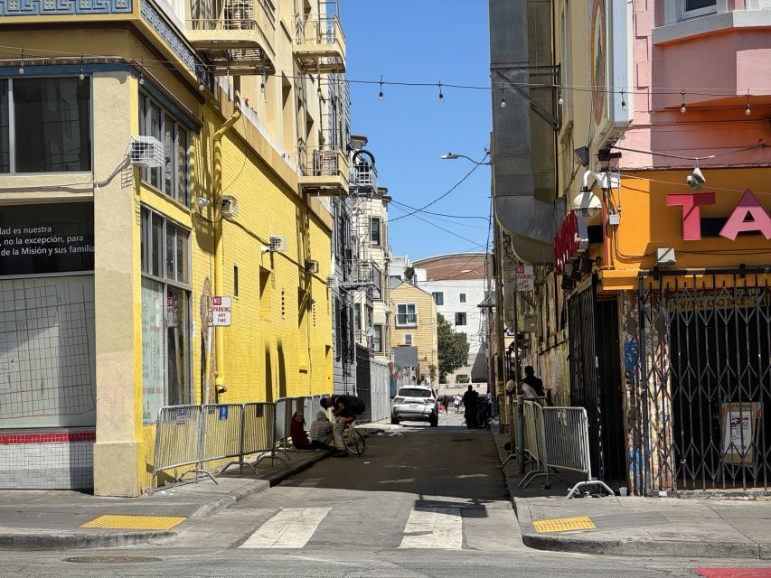 A narrow urban alley with yellow and orange buildings on either side, people sitting on the sidewalk, metal barricades, and a car parked at the end of the alley.
