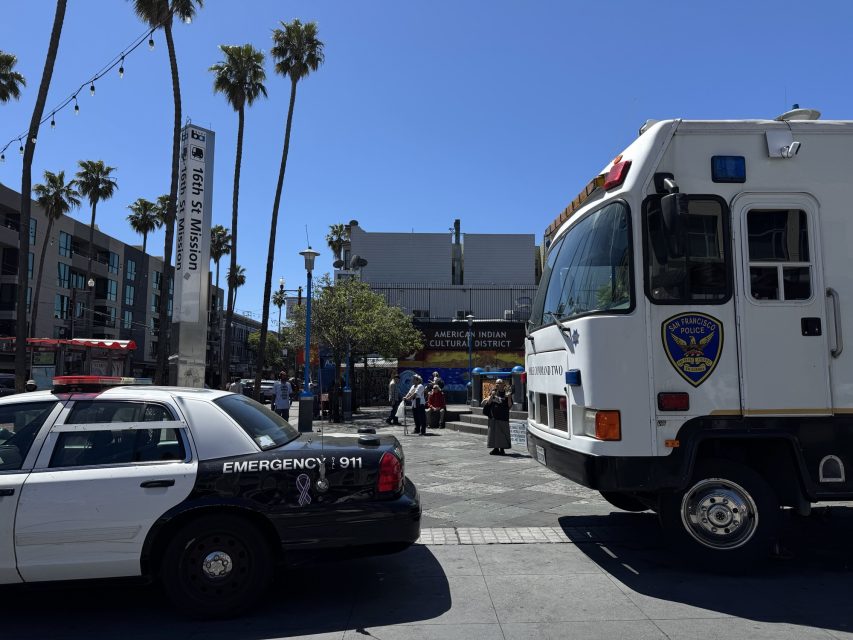Police vehicles are parked in a public square near a sign reading "American Indian Cultural District" under a clear, sunny sky with palm trees and people in the background.