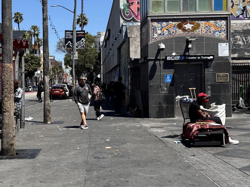 A person sits on a chair with belongings on a city sidewalk while others walk and gather near a building with a BART station entrance.