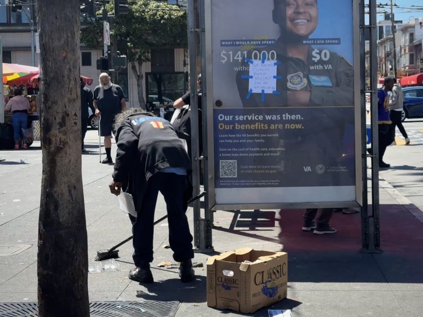 A person sweeps the sidewalk near a bus stop advertisement featuring a veteran. A tree and a cardboard box are also visible in the scene.