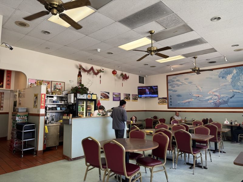 A restaurant with red chairs, round tables, wall art, and ceiling fans. Several people are seated and staff are behind the front counter near a TV and decorative items.