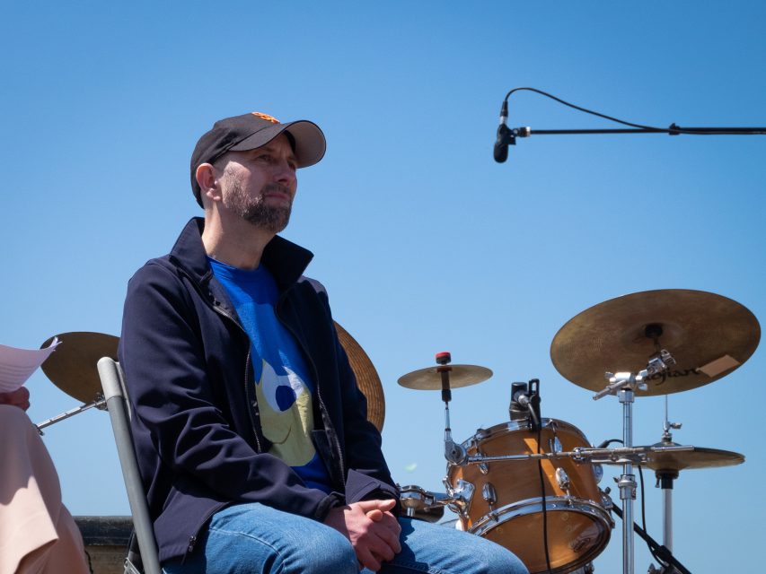 A man wearing a dark jacket and cap sits on a chair outdoors near a drum set, with a large microphone overhead against a clear blue sky.