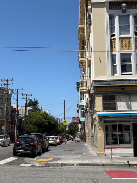 A city street with parked cars, a yellow hotel sign, a crosswalk, power lines, and a beige building under a clear blue sky.