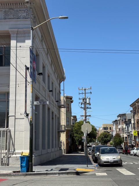 A city street corner with parked cars, a large white building, street signs for 16th St, a utility pole, and clear blue sky.
