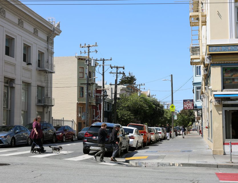 People walk dogs across a city street intersection lined with parked cars and apartment buildings on a clear day.