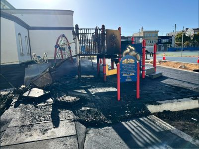 A playground structure and surrounding area appear damaged and partially burned, with melted equipment and dark scorch marks on the ground near a welcome sign.