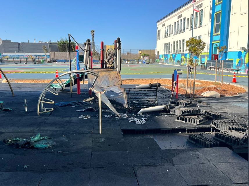 A burned playground structure with melted and charred equipment sits on damaged rubber tiles near a school building on a clear day.