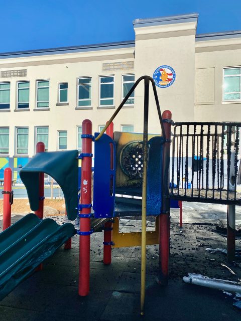 A playground structure with a burnt slide and climbing area, surrounded by debris, is in front of a beige school building.