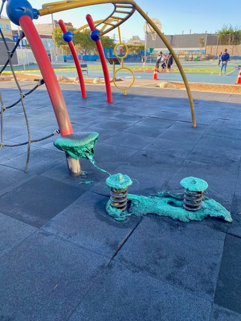 Green foam-like substance oozes from playground equipment onto rubber flooring at an outdoor playground, with other play structures and people visible in the background.