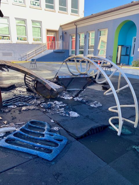 A burnt and partially melted playground structure sits on a damaged rubber surface in front of a school building.
