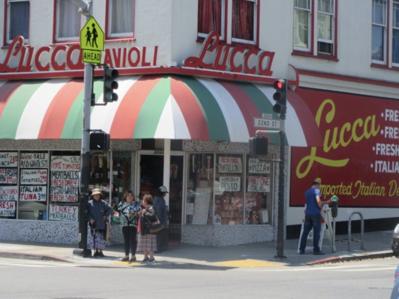 Street view of Lucca Ravioli Company, an Italian deli with red and green striped awning, people standing outside, and hand-painted signs in the windows.