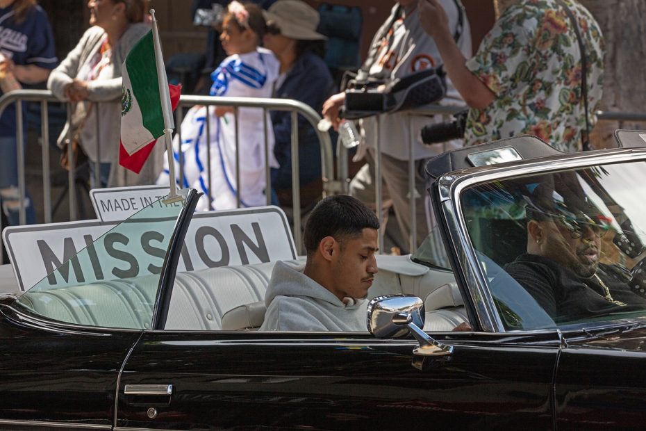 A young man sits in a black convertible with a "MISSION" sign and a Mexican flag in the back, during a street event with people watching behind a barricade.