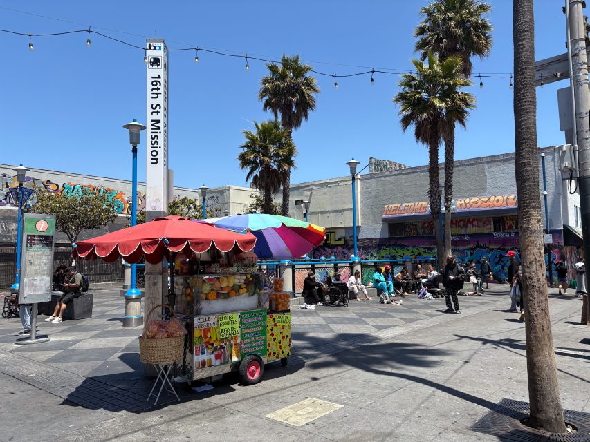 A colorful street food cart with an umbrella stands near the 16th St Mission BART station; people sit and gather nearby, with palm trees and graffiti-covered buildings in the background.