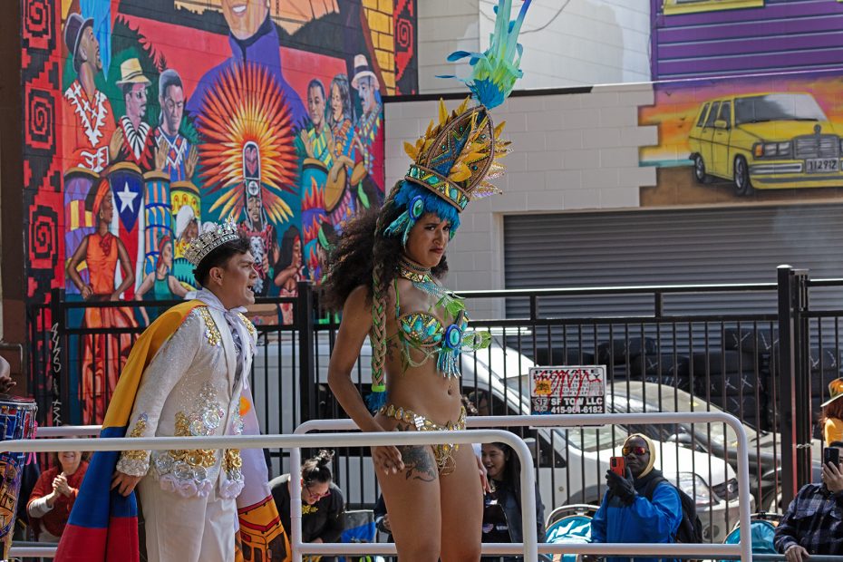 Two people in elaborate costumes participate in a street parade; one wears a feathered headdress and beaded outfit, the other a jeweled jacket and crown, with murals visible in the background.