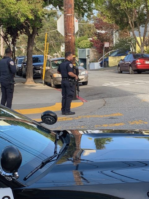 Two police officers stand on a street corner, one holding a rifle. Police vehicles are parked nearby, and the area is cordoned off with yellow tape.