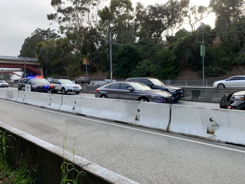 Several cars, including police vehicles with flashing lights, are stopped on a freeway next to a concrete divider; trees and a bridge are in the background.