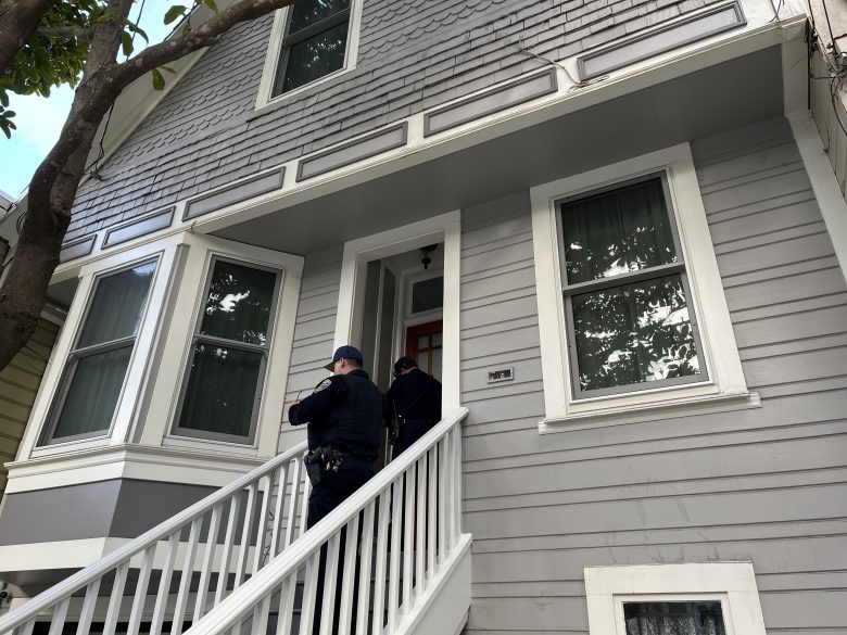 Two police officers stand on the steps of a gray, two-story house, one knocking on the front door.