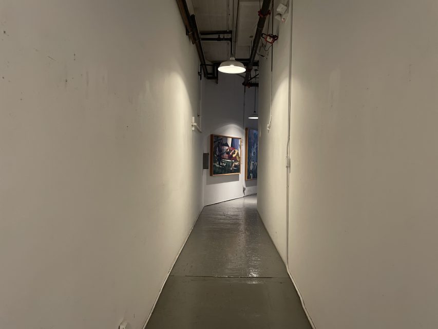 A narrow, dimly lit hallway with white walls and a gray floor, leading to a bend where framed artwork is visible on the right wall. Ceiling lights and exposed pipes overhead.
