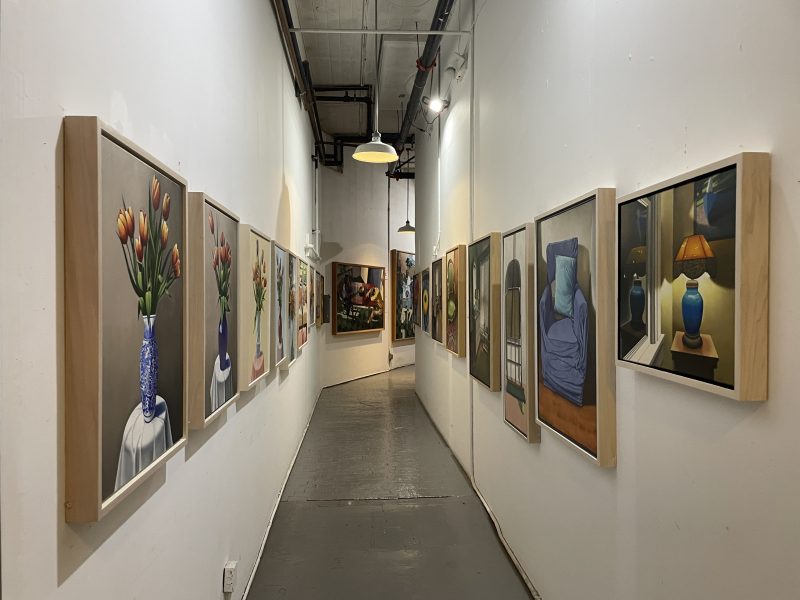 A narrow art gallery hallway with paintings of flowers, vases, and interior scenes displayed on both white walls under ceiling lights.