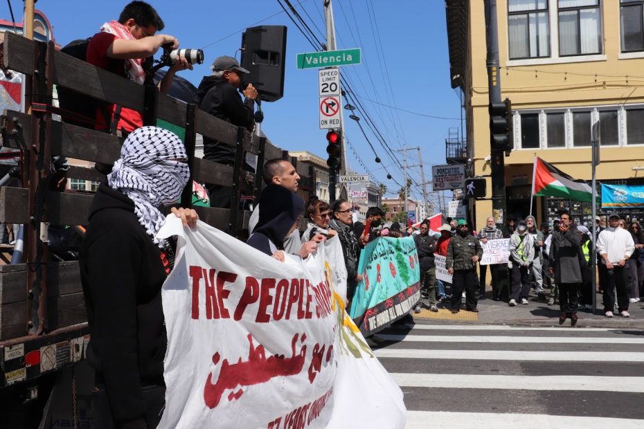 A group of people hold banners and signs at a street protest near the intersection of Valencia Street, with some photographing the event.