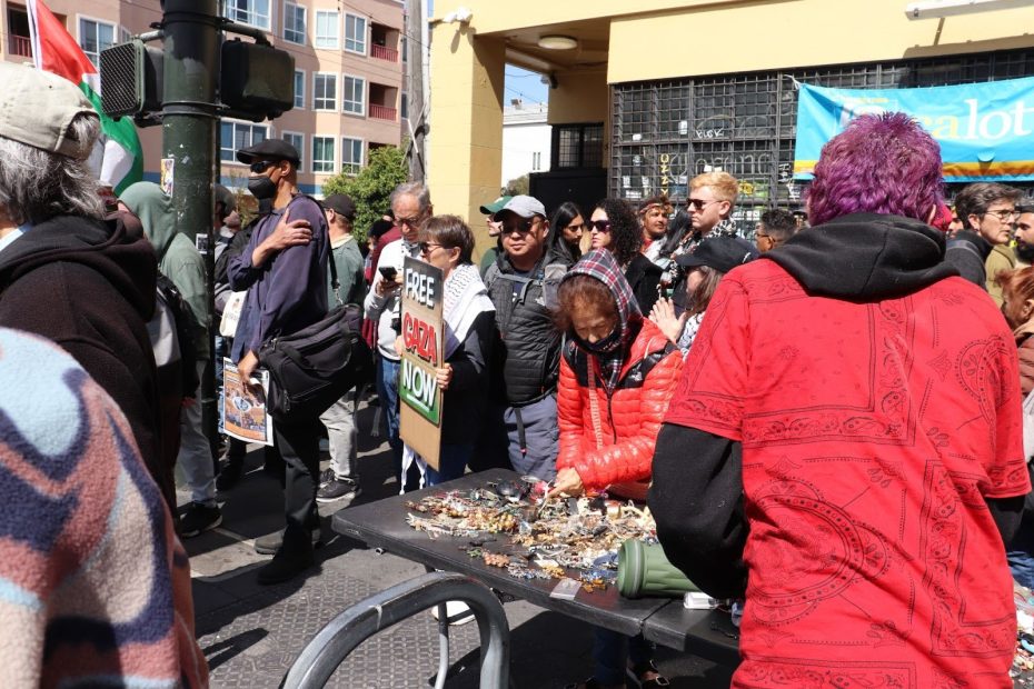 A woman arranges jewelry on a table at an outdoor market while people gather nearby, some holding protest signs and banners.
