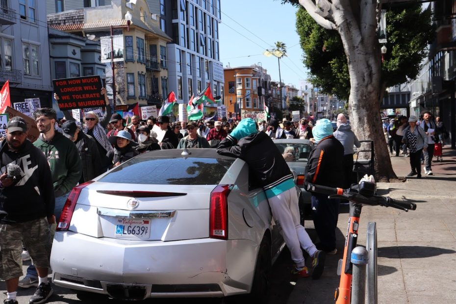 A crowd marches down a city street holding flags and signs, while two people lean over the back of a parked silver Cadillac.