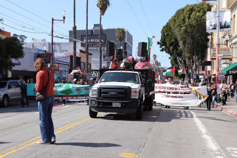 A group of people march down a city street carrying banners, with a pickup truck leading the procession.