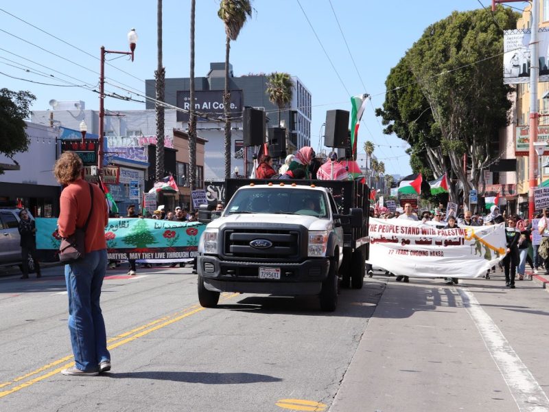 A group of people march down a city street carrying banners, with a pickup truck leading the procession.