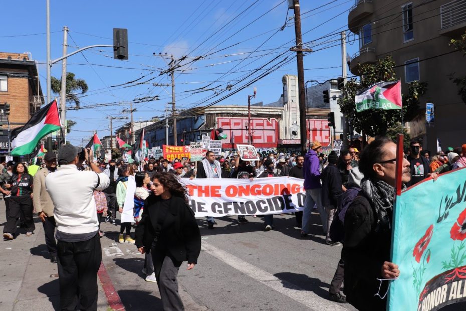A group of people march down a city street holding Palestinian flags and protest signs, including a large banner reading “Stop the ongoing genocide.”.