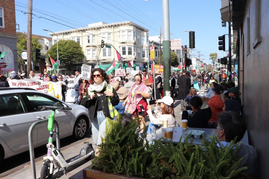 A street protest with people holding signs and flags while others sit at an outdoor cafe; buildings and power lines are visible in the background.