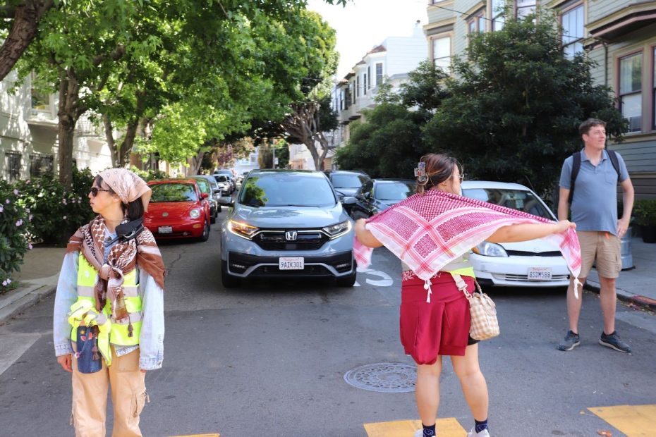 Three people stand in the middle of a residential street with cars waiting behind them; two face away, and one adjusts a scarf.