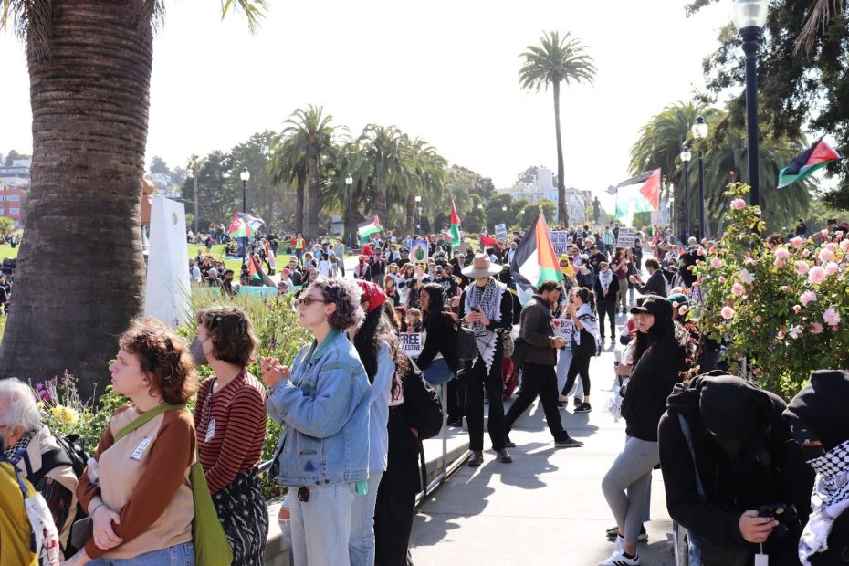 A large group of people gather outdoors in a park, many holding and waving flags, with palm trees and flowers visible in the background.