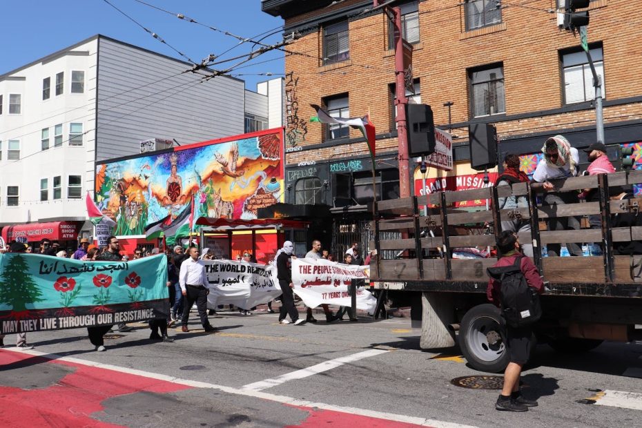 A group of people hold protest banners and flags while walking past a mural and a truck on a city street.