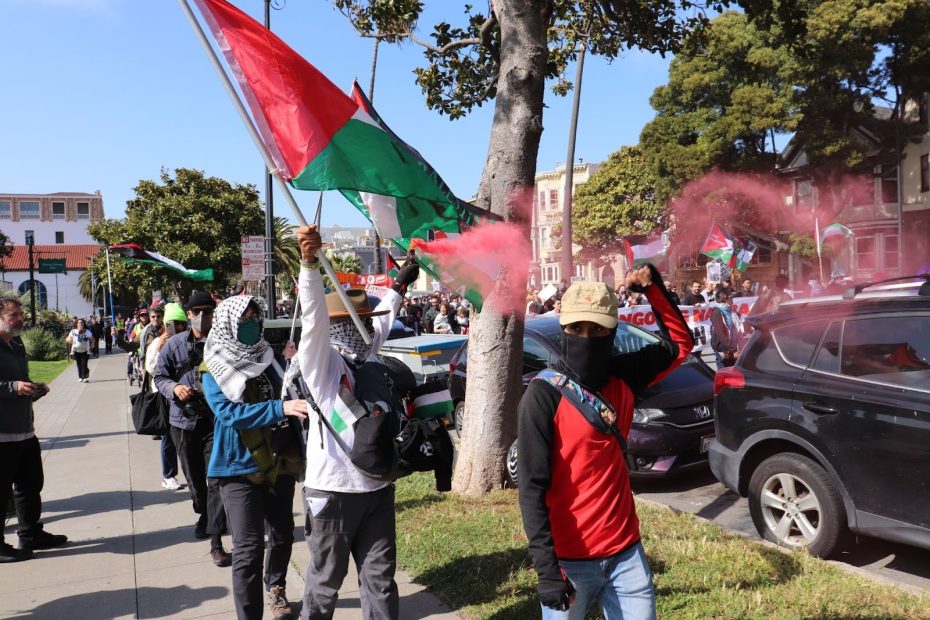 A group of protesters marches on a city sidewalk, holding Palestinian flags and flares, with cars parked nearby and trees in the background.