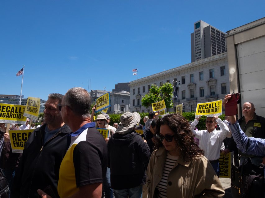 A crowd gathers outside, holding yellow signs that read "RECALL ENGARDIO" in front of city buildings under a clear blue sky.