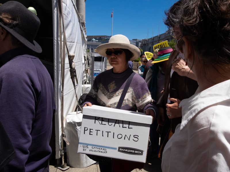 A person wearing sunglasses and a hat holds a white box labeled "RECALL PETITIONS" while standing among a crowd outdoors.