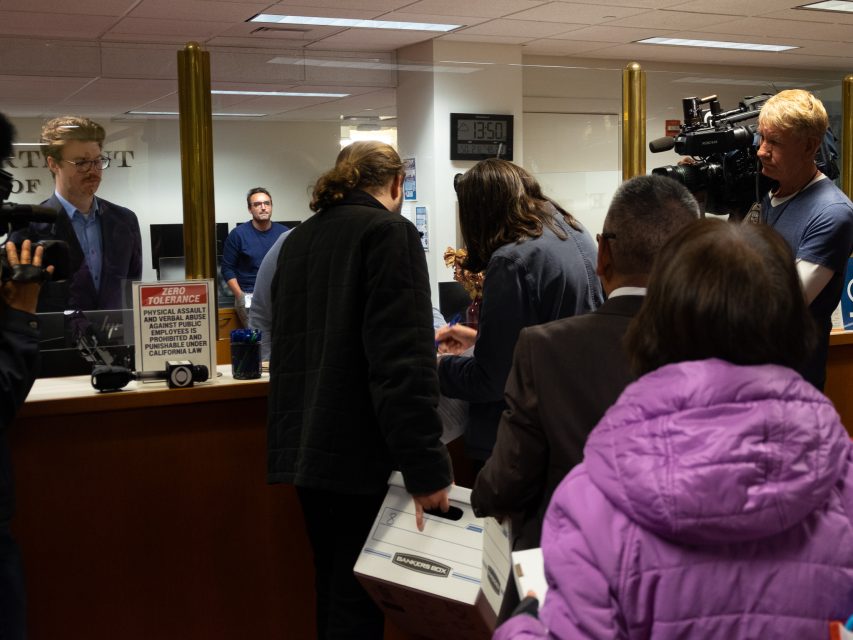 People stand in line at a service counter while being filmed by a camera crew; office staff are visible behind the counter.