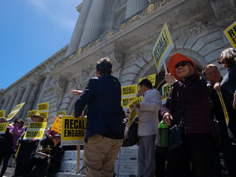 A group of people stand on the steps of a building holding yellow "Recall Engardio" signs during a protest or demonstration.