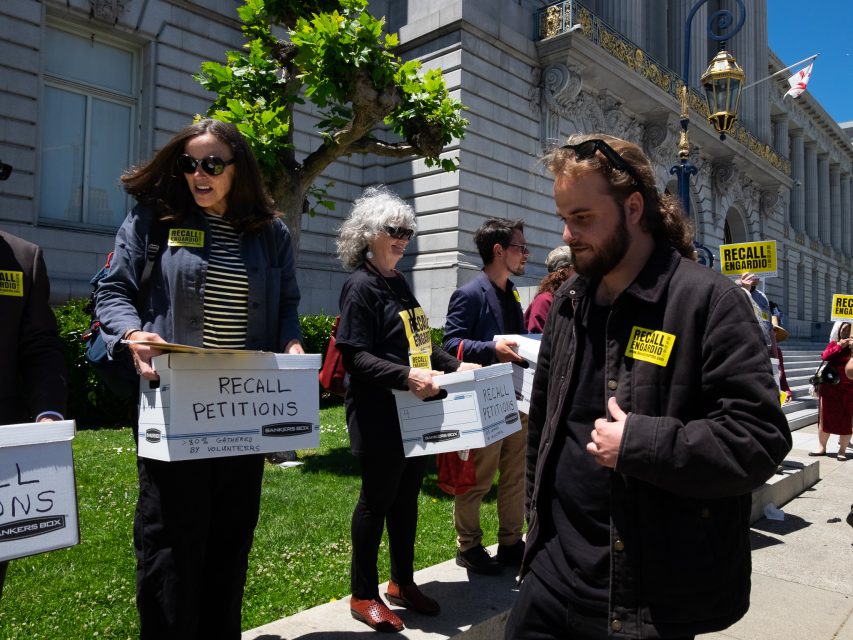 People stand outside a government building holding boxes labeled "Recall Petitions" and wearing jackets with "Recall" stickers, participating in a petition event.