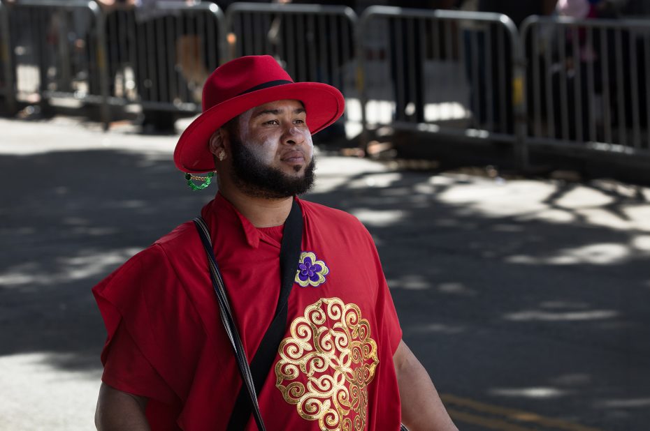 A person wearing a red hat and red clothing with gold patterns walks outdoors, with metal barricades and shadows in the background.