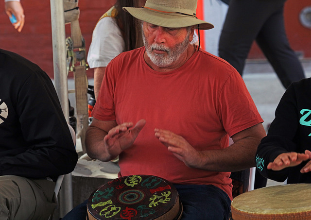 A man in a wide-brim hat and red shirt plays a decorated hand drum, seated beside others, with blurred people in the background.