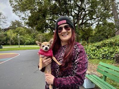 A woman wearing sunglasses and a beanie holds a small dog in a pink sweater, standing on a paved path in a park with trees and a green bench nearby.