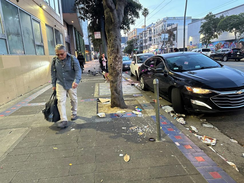 A man carrying a bag walks past a tree and parked cars on a city sidewalk littered with trash and debris.