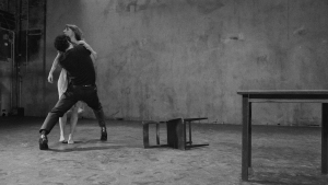 Two people engage in a dramatic physical interaction in a sparse room with a fallen chair and a table against a bare wall.