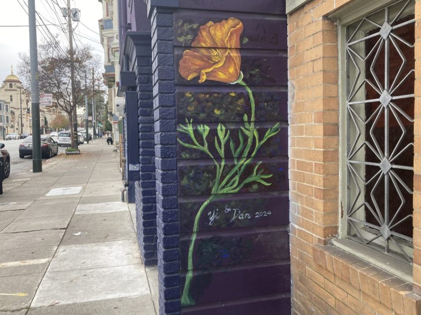 A mural of a large orange-yellow flower with green stem and leaves is painted on a purple brick wall on a city sidewalk. The mural is signed "Jan 2024.